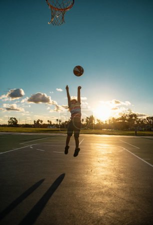 Foto de Lindo niño jugando baloncesto. Lindo niño pequeño saltando con bola de cesta para silueta de tiro en la puesta del sol - Imagen libre de derechos