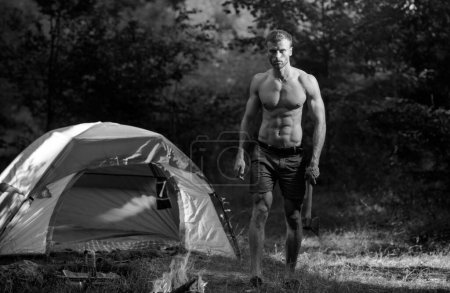 Photo for Handsome and muscular lumberjack man with an axe, camping. Man with axe look at bonfire flame on natural landscape. Guy by a fire in the forest. Traveling, camping hiking vacation - Royalty Free Image