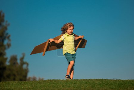 Kid boy playing with cardboard wings. Child in summer field. Kids travel and vacation concept. Imagination and freedom concept. Boy with wings at field imagines pilot and dreams of flying