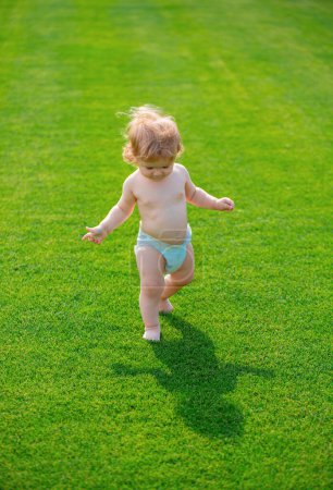 Photo for First step. Baby boy toddler walking in a park on bright spring day. Baby bare legs standing on green grass. Child development - Royalty Free Image