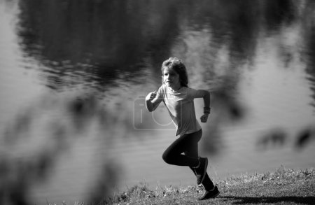 Téléchargez les photos : Enfants courir ou faire du jogging près du lac sur l'herbe dans le parc. Enfant sportif garçon coureur courir dans le parc d'été. Des enfants actifs, des enfants sportifs. Le jogging aide le corps à être fort - en image libre de droit