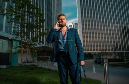 Foto de Retrato sexy hombre de negocios utilizando el teléfono inteligente cerca de edificio moderno al aire libre. Hombre de negocios exitoso caminando en traje en la calle - Imagen libre de derechos
