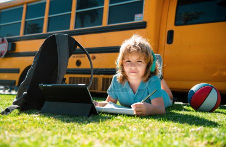 Téléchargez les photos : L'enfant élève fait ses devoirs à l'école en s'allongeant sur l'herbe dans le parc près de l'autobus scolaire. École enfant en plein air - en image libre de droit