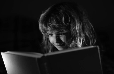Téléchargez les photos : Garçon lisant l'histoire du coucher, photo de chambre noire. Enfant lisant un livre magique au lit. Enfant garçon lire des livres à la maison - en image libre de droit