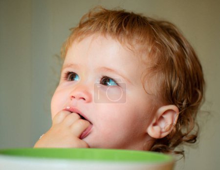 Téléchargez les photos : Bébé drôle mangeant lui-même avec une cuillère sur la cuisine. Lèche de savoureux doigts. Enfant drôle visage gros plan - en image libre de droit