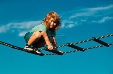 Téléchargez les photos : Mignon garçon monte l'échelle sur l'aire de jeux. L'enfant monte l'échelle contre le ciel bleu. Beau garçon mignon souriant sur une aire de jeux. Activités pour enfants - en image libre de droit