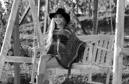 Foto de Joven mujer sexy en estilo boho en el sombrero se balancea en un columpio en el jardín del parque de verano. Chica rubia sensual descansando en el salón. Chica Hipster - Imagen libre de derechos