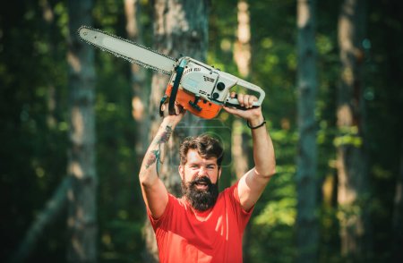 Photo for Lumberjack worker walking in the forest with chainsaw. Firewood as a renewable energy source. A handsome young man with a beard carries a tree - Royalty Free Image