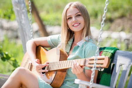 Photo for Young Woman sit on swing and playing guitar in summer park. Blonde Female model with guitar. Girl playing guitar outdoor. Singing Girl With Guitar. Summer music - Royalty Free Image
