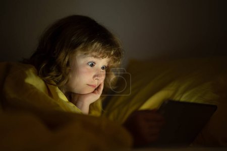 Foto de Cuento para dormir. Niño viendo la tableta antes de dormir. El niño lee el libro electrónico. El niño mira la pantalla de la tableta a la altura. Niño jugando tableta acostado en una cama debajo de la manta - Imagen libre de derechos