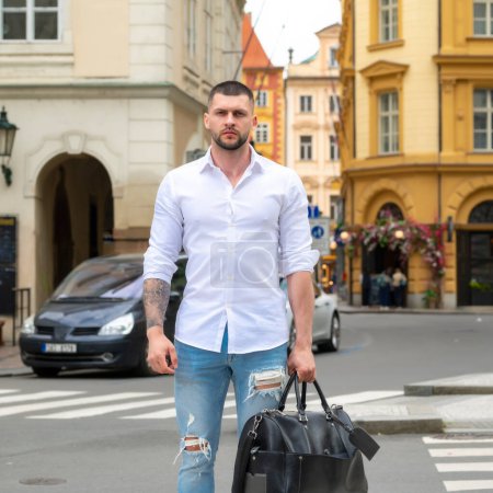 Téléchargez les photos : Beau jeune homme en voyage d'affaires marchant avec ses bagages dans la rue. Homme d'affaires itinérant - en image libre de droit
