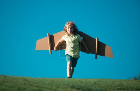 Téléchargez les photos : Un gamin jouant avec des ailes en carton. Enfant dans un champ d'été. Les enfants voyagent et vacances concept. Concept d'imagination et de liberté - en image libre de droit