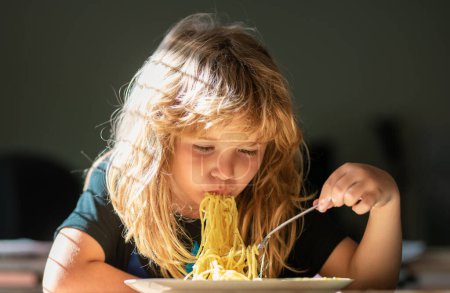 Téléchargez les photos : Un gamin qui mange des spaghettis dans la cuisine. Jeune enfant assis sur la table manger des aliments sains avec une expression drôle sur le visage. Gros plan portrait de gosse drôle manger des nouilles pâtes - en image libre de droit