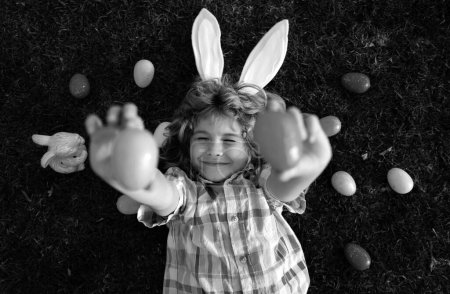 Foto de Conejito de Pascua niño cazando huevos al aire libre Feliz día de Pascua, Niño que pone en la hierba en el parque. Top view niños divertidos que ponen en la hierba - Imagen libre de derechos