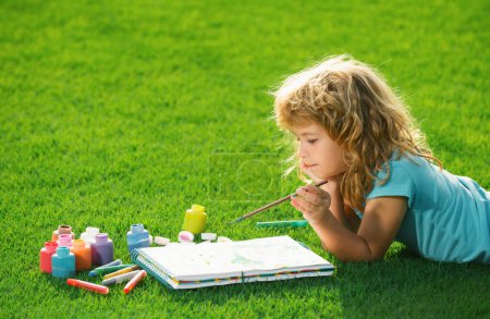 Foto de Pintura infantil dibujo arte. Niño niño dibuja en parque tendido en la hierba divertirse en el fondo de la naturaleza - Imagen libre de derechos