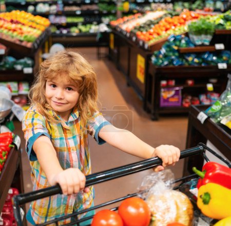 Foto de Little cute boy with shopping cart full of fresh organic vegetables and fruits in grocery food store or supermarket. Boy having fun while choosing food in the supermarket - Imagen libre de derechos