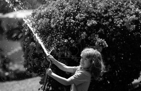 Foto de Lindo niño regando flores en el jardín en el día de verano. Niño agricultor con manguera de jardín en planta de plantación. Manguera. Divertido niño regando plantas en el jardín del patio - Imagen libre de derechos