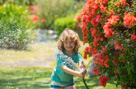 Téléchargez les photos : Mignon garçon arrosant des plantes dans le jardin le jour de l'été. Enfant avec outils de jardin et tuyau d'arrosage dans le jardin arrière-cour. Le gamin s'amuse dans la cour. Le concept de gentillesse et d'enfance des enfants - en image libre de droit