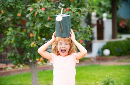 Téléchargez les photos : Amusant enfant étonné arrosant des arbres et des fleurs dans le jardin familial à une journée d'été. Un gamin aide à la cour - en image libre de droit