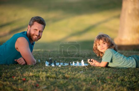 Téléchargez les photos : Père et fils jouant aux échecs allongés sur l'herbe à Lawn Park. Fête des Pères, famille amoureuse, parentalité, concept d'enfance. Intelligent penser enfant tout en jouant aux échecs - en image libre de droit