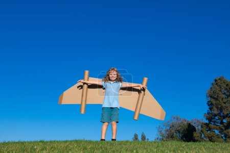 Téléchargez les photos : Joyeux enfant jouant avec des ailes de jouet sur fond de ciel d'été. Succès des enfants, concept de leader et gagnant - en image libre de droit