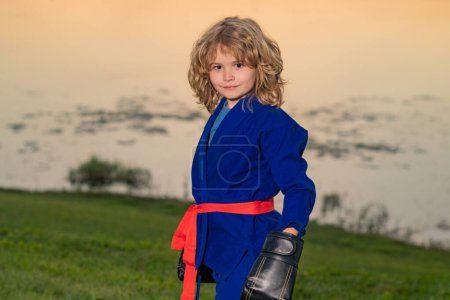 Photo for Kid boy practicing box outdoor. Sport karate kids. Little boy in kimono doing box in park. Child with boxing gloves training box. Little fighter. Martial arts kids. Child in kimono doing boxing punch - Royalty Free Image