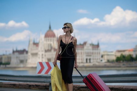 Foto de Viaje a Europa. Mujer turista en vestido de moda con maleta caminando por la calle. Chica estudiante viajero retrato al aire libre. Jóvenes turistas caminando por la calle de la ciudad - Imagen libre de derechos