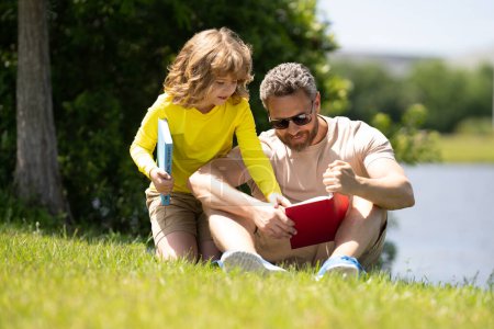 Téléchargez les photos : Père et fils assis sur l'herbe verte dans le jardin et le livre de lecture ensemble. Joyeux livre de lecture en famille ensemble dans la nature verte. Père est assis avec son fils lire le livre. Le gamin est très intéressé. - en image libre de droit
