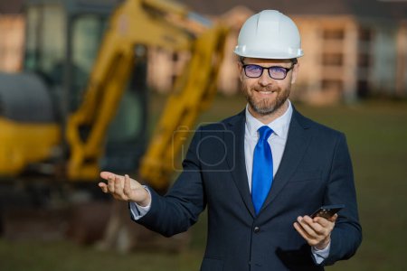 Photo for Civil engineer worker at a construction site. Mature engineer worker. Man in suit and hardhat helmet at construction site. Middle aged head civil engineer worker standing outside near excavator - Royalty Free Image
