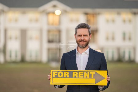 Photo for Realtor agent is a realtor with sign for rent in hand against the background on new apartment home background. Realtor in suit, outdoor portrait. Realtor renting new home. Property rental concept - Royalty Free Image