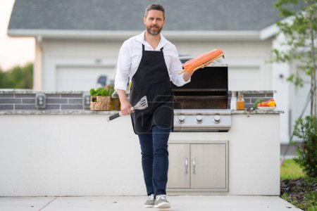 Barbecue master in chef apron hold salmon fillet on BBQ. Millennial man in apron for barbecue. Roasting and grilling salmon fillet. Man hold cooking utensils barbecue. Roasting salmon fish