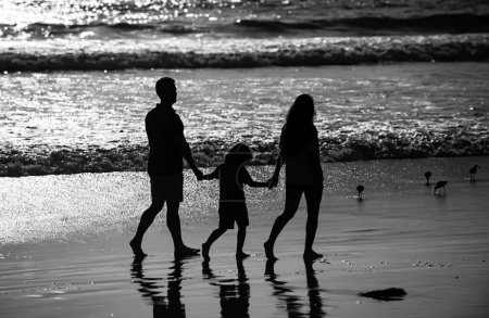Foto de Silueta de familia caminando por la playa. Padres y niños caminando sobre arena dorada, mamá y papá tomados de las manos de las niñas. Longitud total. Familia actividades al aire libre concep - Imagen libre de derechos
