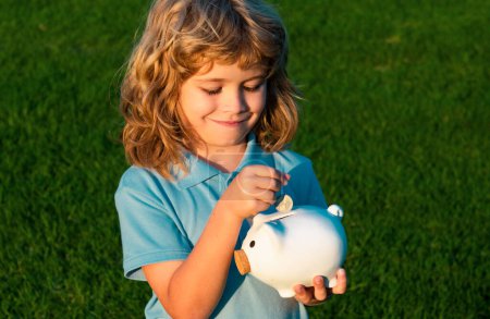 Photo for Child boy putting in piggy bank coin money. Portrait of little boy with moneybox piggybank on grass background - Royalty Free Image