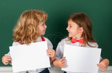 Photo for Schoolkids holding white paper blank, poster with copy space. Happy girl and boy school friends. Face portrait of two schoolkids. Children couple at primary school classmates, young students - Royalty Free Image