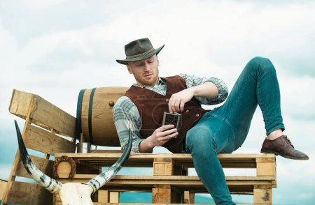 Téléchargez les photos : Un cow-boy fermier portant un chapeau. La vie occidentale. Portrait masculin country américain. Exploitant agricole à la campagne à la ferme ou au ranch. Homme attrayant avec whisky ou brandy - en image libre de droit