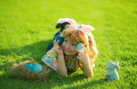 Cute child bunny wear rabbit ears laying on grass in park, Happy Easter day. Bunny boy, funny outdoor portrait. Hunting eggs and spring holidays with children