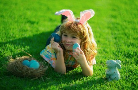 Photo for Child with rabbit ears, Easter holidays. Kid hunting Easter eggs. Cute bunny boy laying on grass in park, funny kids portrait - Royalty Free Image