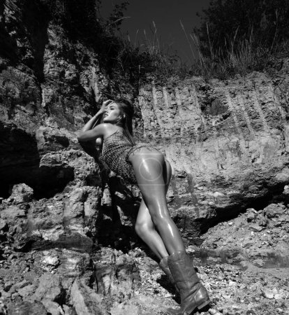 Foto de Mujer sexy cuerpo en las rocas al aire libre, de cuerpo entero. Mujer de moda elegante posando al aire libre. Atractiva joven mujer en vestido sexy disfrutando del verano. Botas occidentales - Imagen libre de derechos