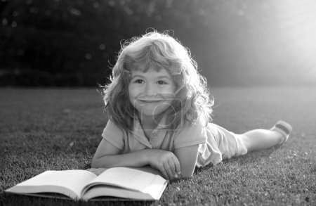 Foto de Niños inteligentes. Lindo chico leyendo un libro tendido en la hierba. Niño leyendo un libro en el parque de verano - Imagen libre de derechos