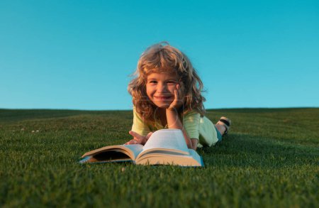 Foto de Niño inteligente leyendo libro en el parque al aire libre. Sonriente niño divertido en camiseta que se divierte leyendo libro en el parque - Imagen libre de derechos