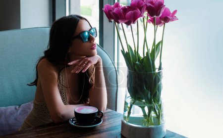 Téléchargez les photos : Femme romantique avec tasse de café. Jeune femme élégante, lunettes de soleil de mode, assis dans un café avec cappuccino tasse. Tulipes de printemps - en image libre de droit