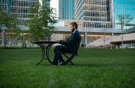 Téléchargez les photos : Homme d'affaires d'âge moyen tapant sur ordinateur portable assis à table sur la terrasse du bureau. Homme travaillant à distance dans un parc moderne - en image libre de droit