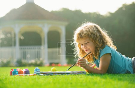 Téléchargez les photos : Peinture d'enfant dessin art. Kid dessine dans le parc posé dans l'herbe en s'amusant sur fond de nature. Cours de peinture à l'école, dessin d'art - en image libre de droit