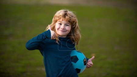 Photo for Boy child playing football on football field. Kid playing soccer show thumbs up success sign. Boy holding soccer ball, close up sporty kids portrait - Royalty Free Image
