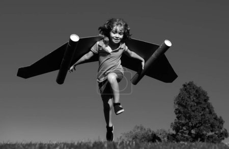 Téléchargez les photos : Joyeux enfant courant avec des ailes en papier sautant contre le ciel bleu. Portrait de garçon jouant avec jetpack jouet. Liberté insouciante, gagnant de succès, concept d'innovation et d'imagination - en image libre de droit