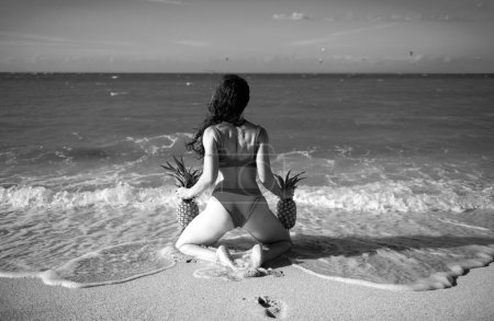 Téléchargez les photos : Belle fille modèle sexy en bikini au bord de la mer de l'île tropicale. Voyage vacances d'été. Femme sexy tenant des ananas, vue sur le dos - en image libre de droit
