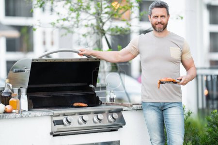 Photo for Man at barbecue grill. Male cook preparing barbecue outdoors. Bbq meat, grill for picnic. Roasted on barbecue. Man preparing barbeque in the house yard. Barbecue and grill. Cook using barbeque tongs - Royalty Free Image
