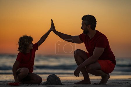 Photo for Father and son sitting on the beach of summer with sunset tone. Child and father building sandcastle. Father and son playing on summer beach. Father and child son playing with sand on sea. Five hand - Royalty Free Image