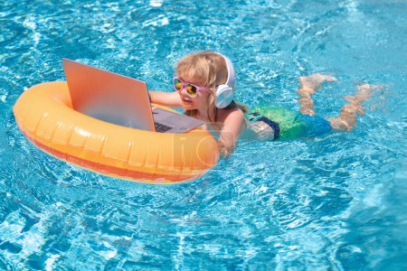 Téléchargez les photos : Ordinateur portable dans la piscine. Enfant assis dans l'anneau de natation dans la piscine et en utilisant un ordinateur portable. Shopping en ligne, concept freelance, voyage d'été - en image libre de droit