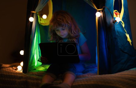 Téléchargez les photos : Enfant garçon couché au lit et regardant ordinateur portable la nuit. Petit hacker, jeune programmeur - en image libre de droit